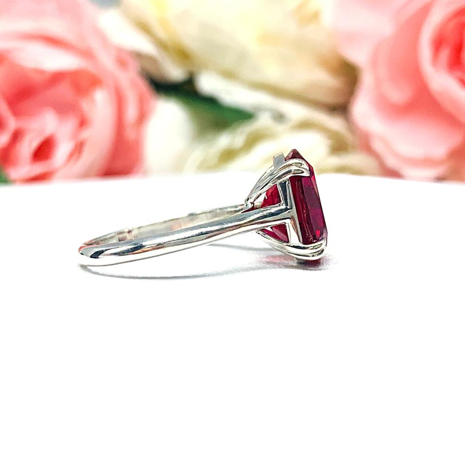 Sterling silver ring featuring a vivid elongated cushion-cut lab created ruby held in a split-prong setting, with soft-focus pink and white flowers in the background and “SJ” initials subtly visible in the corner.