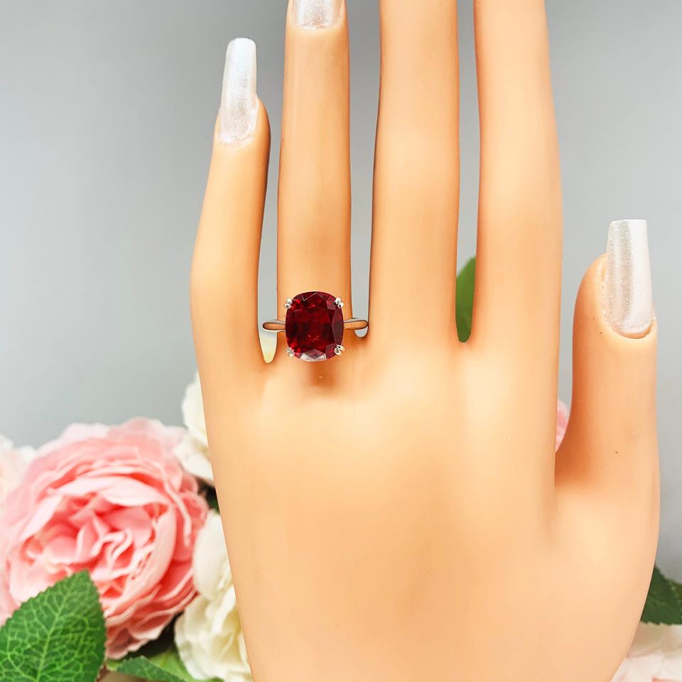 Mannequin hand with long white shimmery nails, displayed against a soft white-gray background with pink and white floral accents. The hand is wearing a sterling silver ring featuring a vivid elongated cushion-cut lab created ruby held in a split-prong setting.