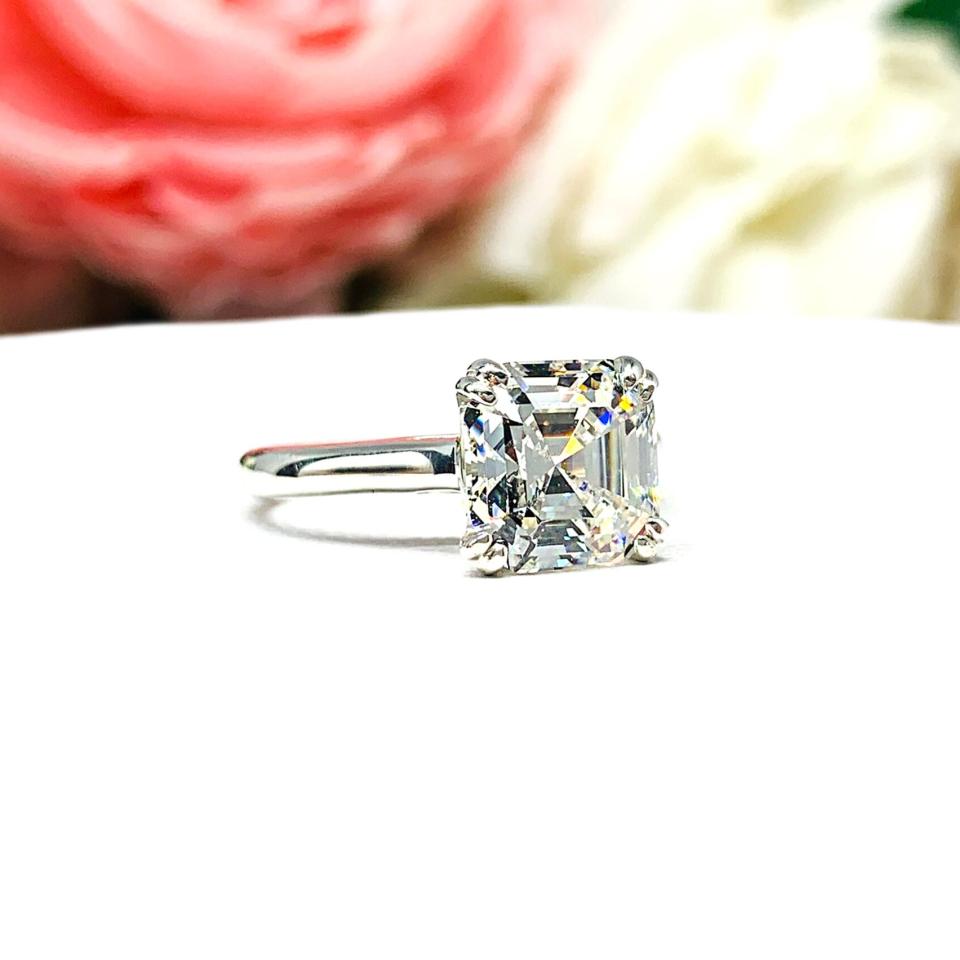 Sterling silver 8mm near colorless (G/H color) asscher cut cubic zirconia solitaire engagement ring. The ring is resting on a white surface with a background of pink and white flowers.