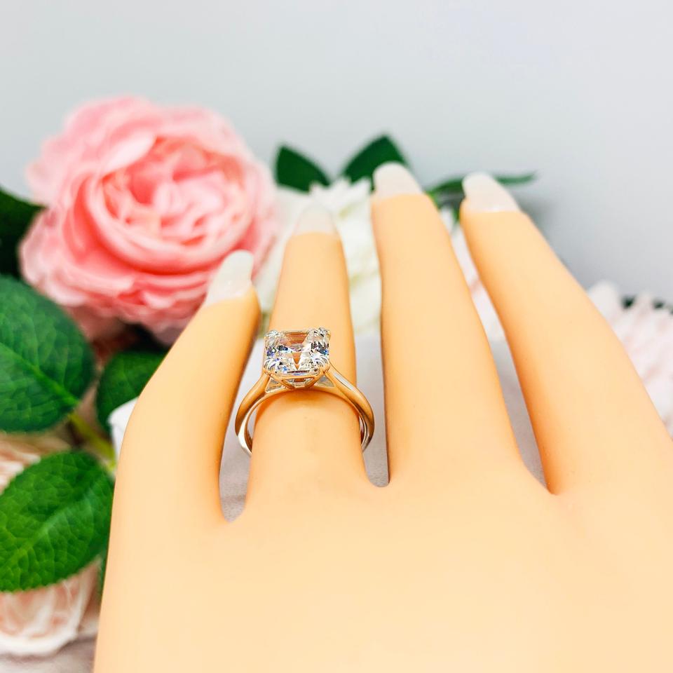 Mannequin hand with long white shimmery nails, displayed against a soft white-gray background with pink and white floral accents. The hand is wearing a sterling silver 8mm near colorless (G/H color) asscher cut cubic zirconia solitaire engagement ring. The ring is resting on a white surface with a background of pink and white flowers.