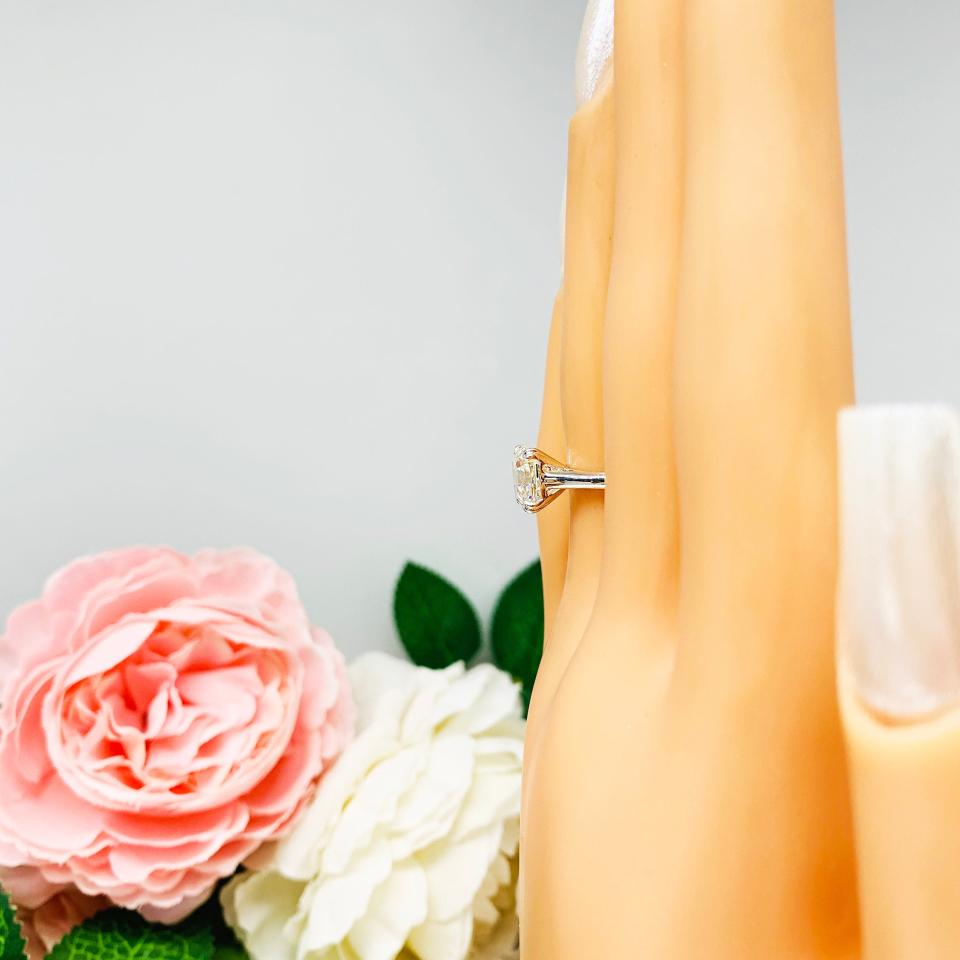 Mannequin hand with long white shimmery nails, displayed against a soft white-gray background with pink and white floral accents. The hand is wearing a sterling silver 8mm near colorless (G/H color) asscher cut cubic zirconia solitaire engagement ring. The ring is resting on a white surface with a background of pink and white flowers.