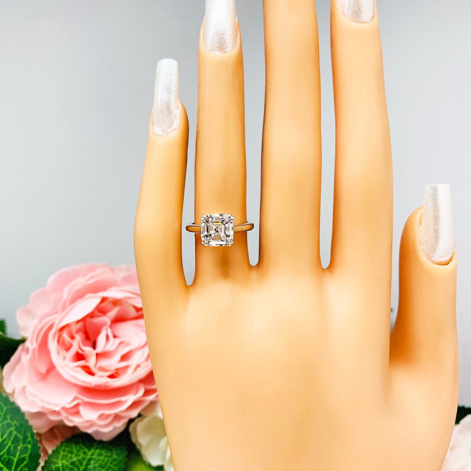 Mannequin hand with long white shimmery nails, displayed against a soft white-gray background with pink and white floral accents. The hand is wearing a sterling silver 8mm near colorless (G/H color) asscher cut cubic zirconia solitaire engagement ring. The ring is resting on a white surface with a background of pink and white flowers.