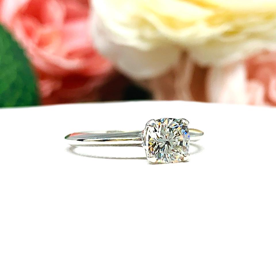 Sterling silver engagement ring set with an 6mm brilliant cushion near colorless cubic zirconia. The ring is resting on a white surface with a background of soft-focus pink and white flowers.