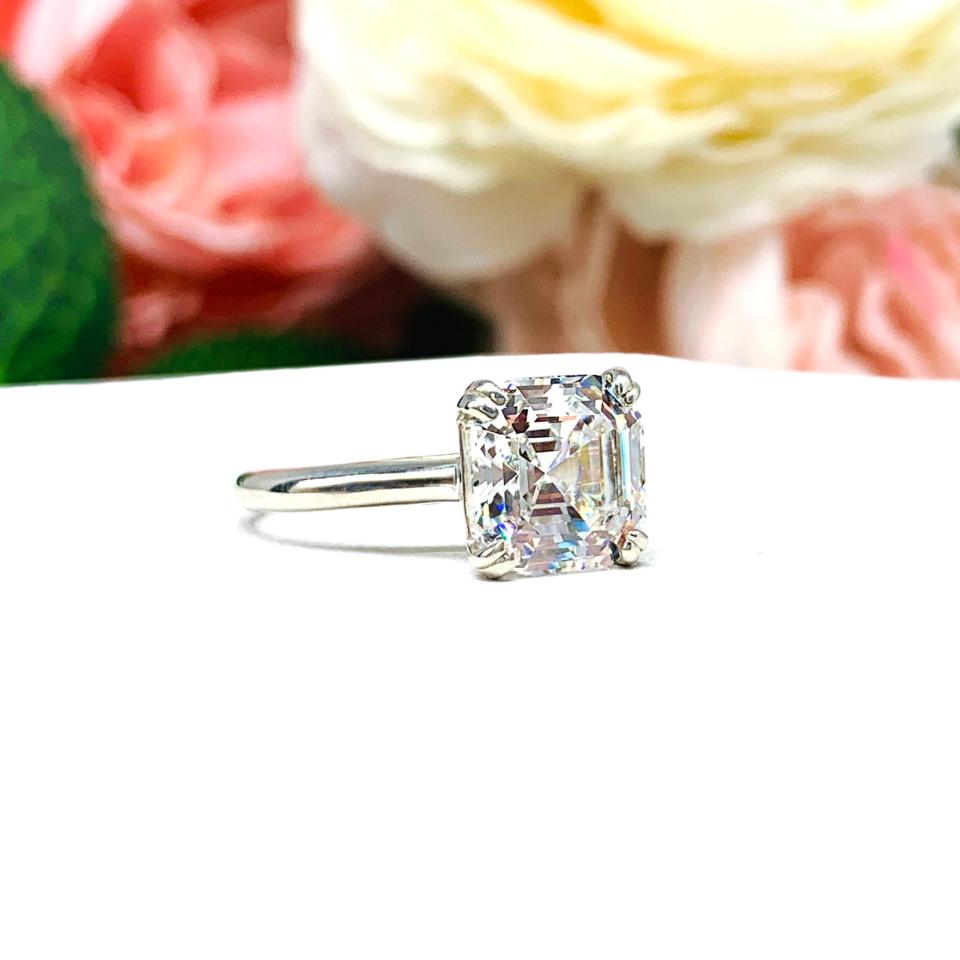 Sterling silver 9mm near colorless (G/H color) asscher cut cubic zirconia solitaire engagement ring. The ring is resting on a white surface with a background of pink and white flowers.