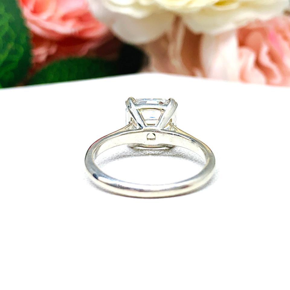 Sterling silver 9mm near colorless (G/H color) asscher cut cubic zirconia solitaire engagement ring. The ring is resting on a white surface with a background of pink and white flowers.