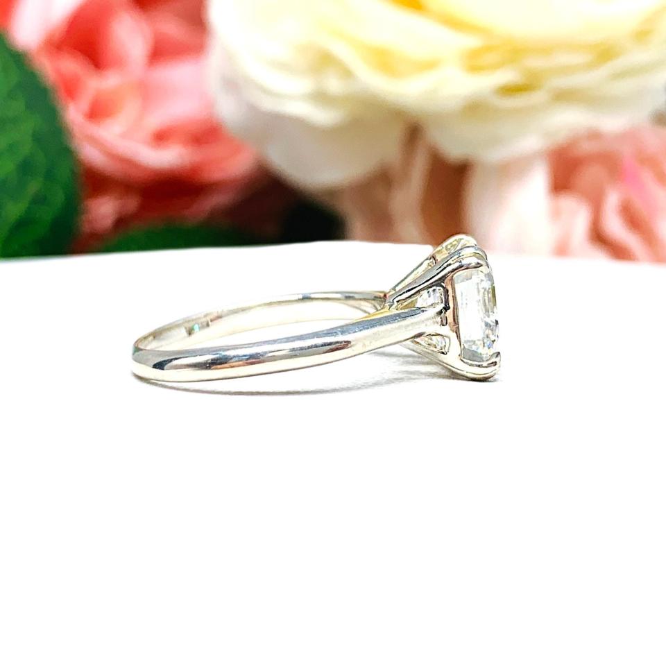 Sterling silver 9mm near colorless (G/H color) asscher cut cubic zirconia solitaire engagement ring. The ring is resting on a white surface with a background of pink and white flowers.