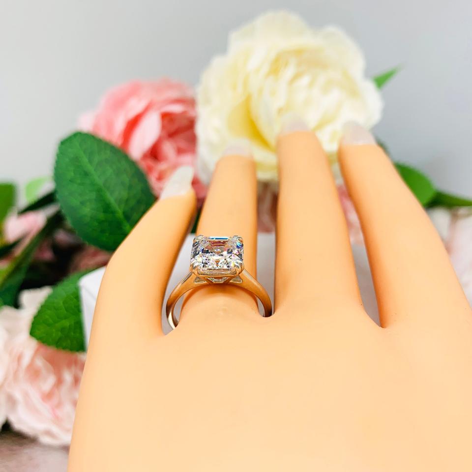 Mannequin hand with long white shimmery nails, displayed against a soft white-gray background with pink and white floral accents. The hand is wearing a sterling silver 9mm near colorless (G/H color) asscher cut cubic zirconia solitaire engagement ring.