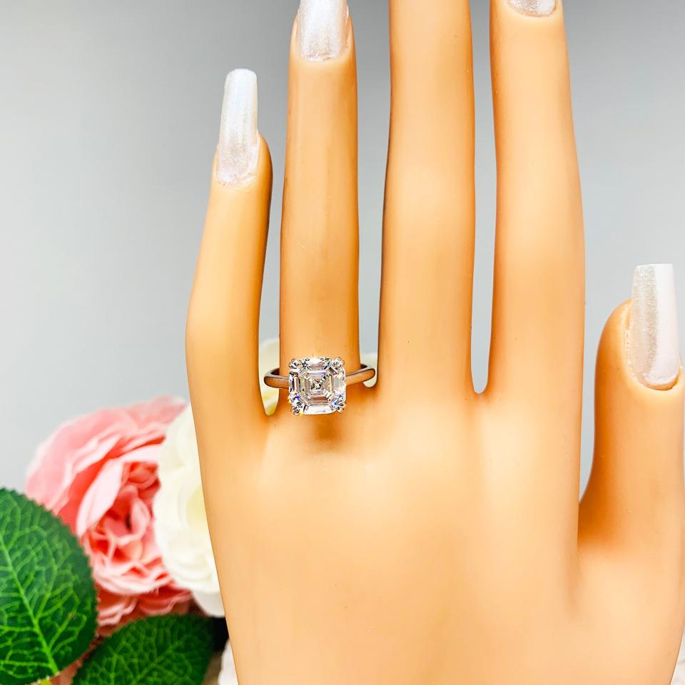Mannequin hand with long white shimmery nails, displayed against a soft white-gray background with pink and white floral accents. The hand is wearing a sterling silver 9mm near colorless (G/H color) asscher cut cubic zirconia solitaire engagement ring.