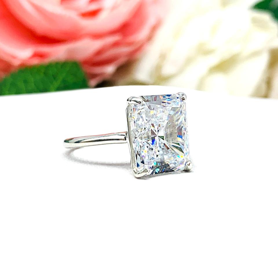 Elegant sterling silver cathedral engagement ring set with a large radiant cut cubic zirconia, beautifully photographed against a romantic and timeless soft-focus floral background of pink and white blooms.