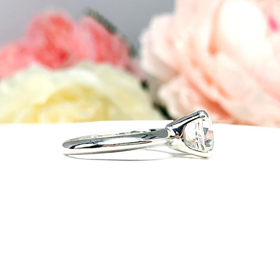 Sterling silver engagement ring set with an 8mm old mine cut near colorless cubic zirconia. The ring is resting on a white surface with a background of soft-focus pink and white flowers.