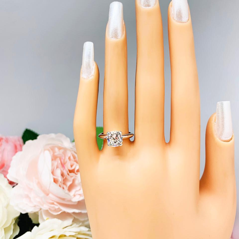 Mannequin hand with long white shimmery nails, displayed against a soft white-gray background with pink and white floral accents. The hand is wearing a sterling silver engagement ring set with an 8mm old mine cut near colorless cubic zirconia.