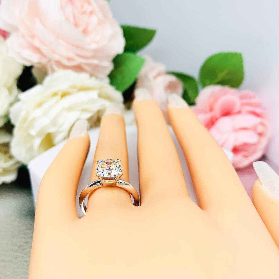Mannequin hand with long white shimmery nails, displayed against a soft white-gray background with pink and white floral accents. The hand is wearing a sterling silver engagement ring set with an 8mm old mine cut near colorless cubic zirconia.
