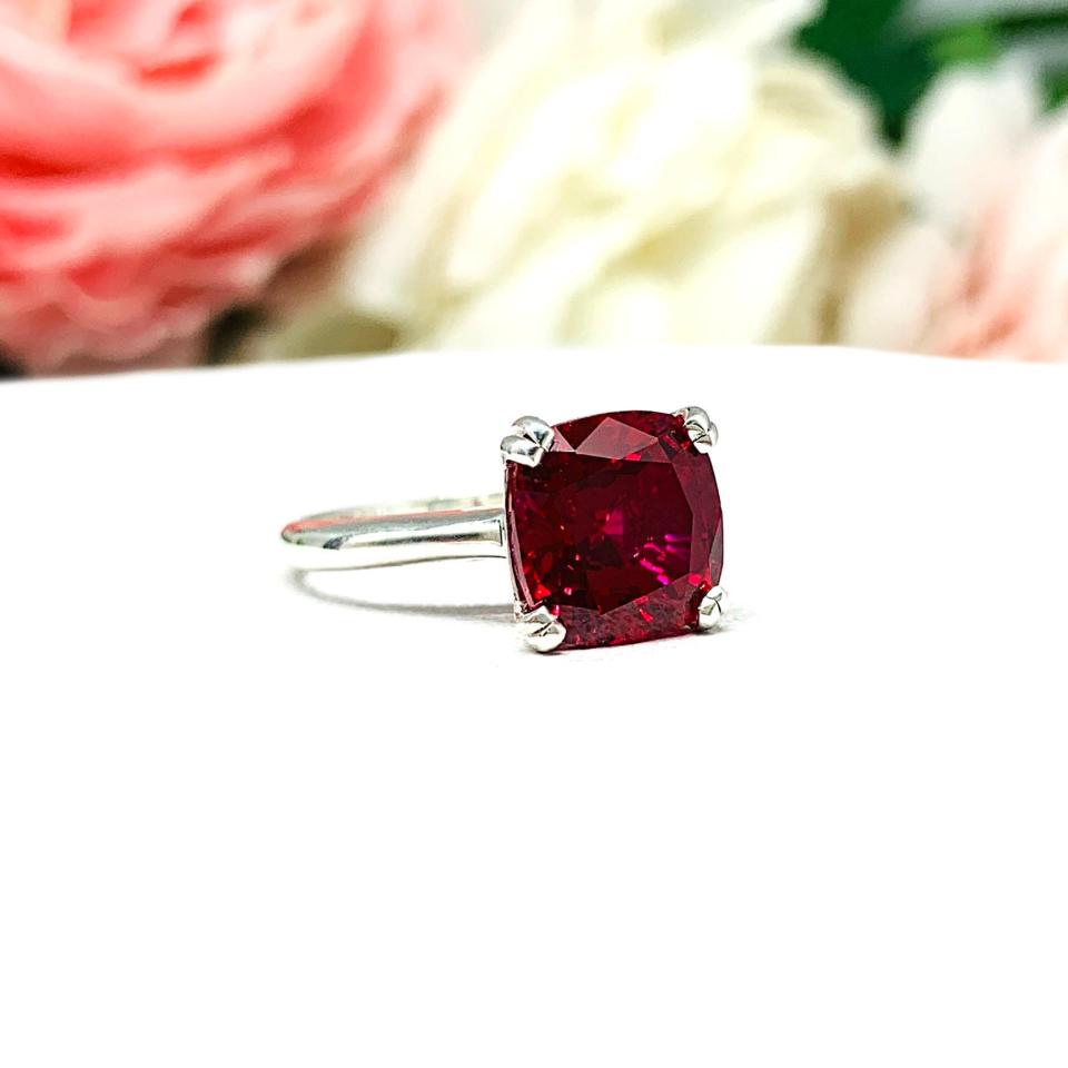 Sterling silver ring with a cushion cut lab created deep red ruby set in a split-prong setting set against a white surface with soft-focus pink and white flowers in the background.
