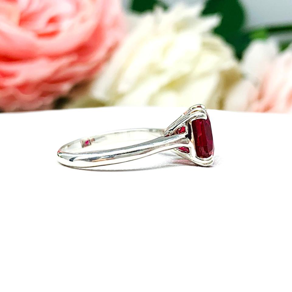 Sterling silver ring with a cushion cut lab created deep red ruby set in a split-prong setting set against a white surface with soft-focus pink and white flowers in the background.