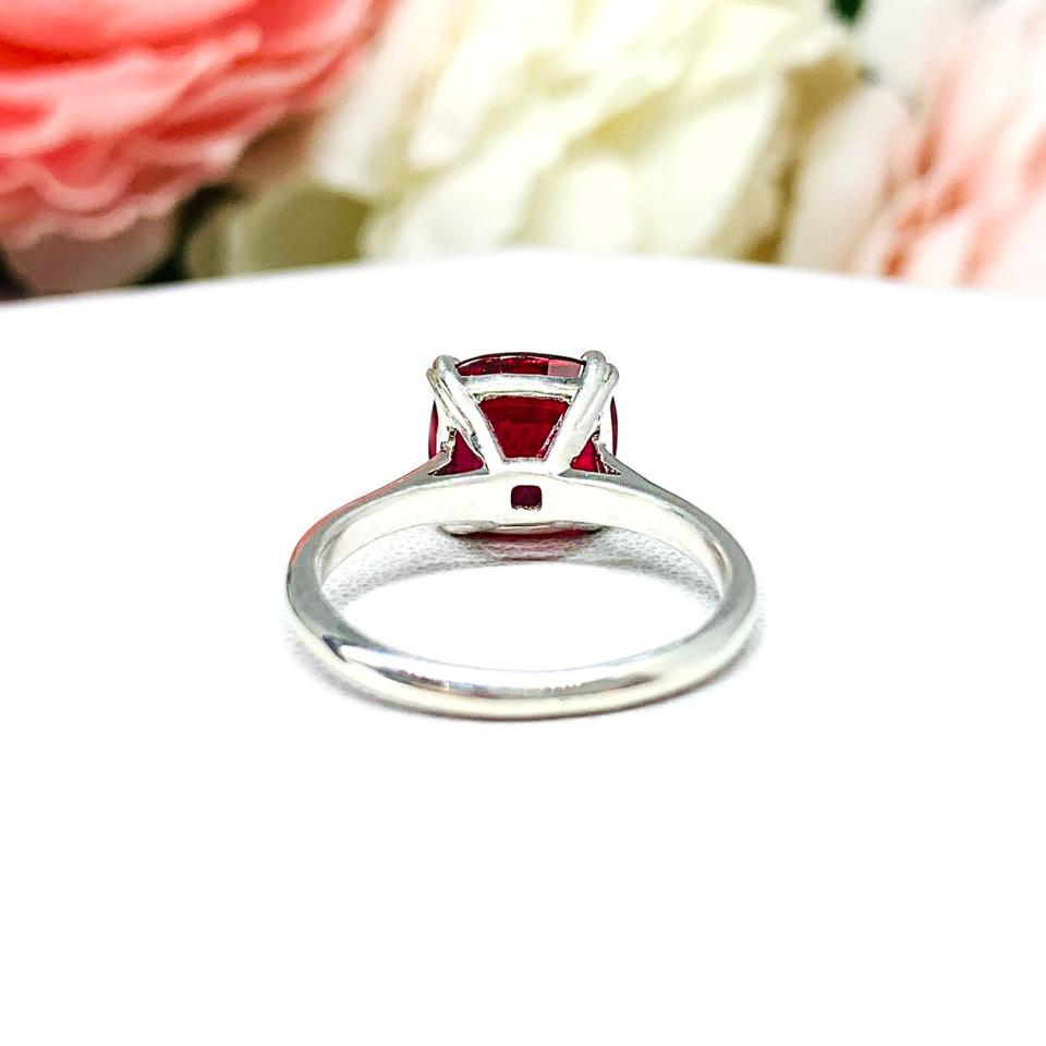 Sterling silver ring with a cushion cut lab created deep red ruby set in a split-prong setting set against a white surface with soft-focus pink and white flowers in the background.