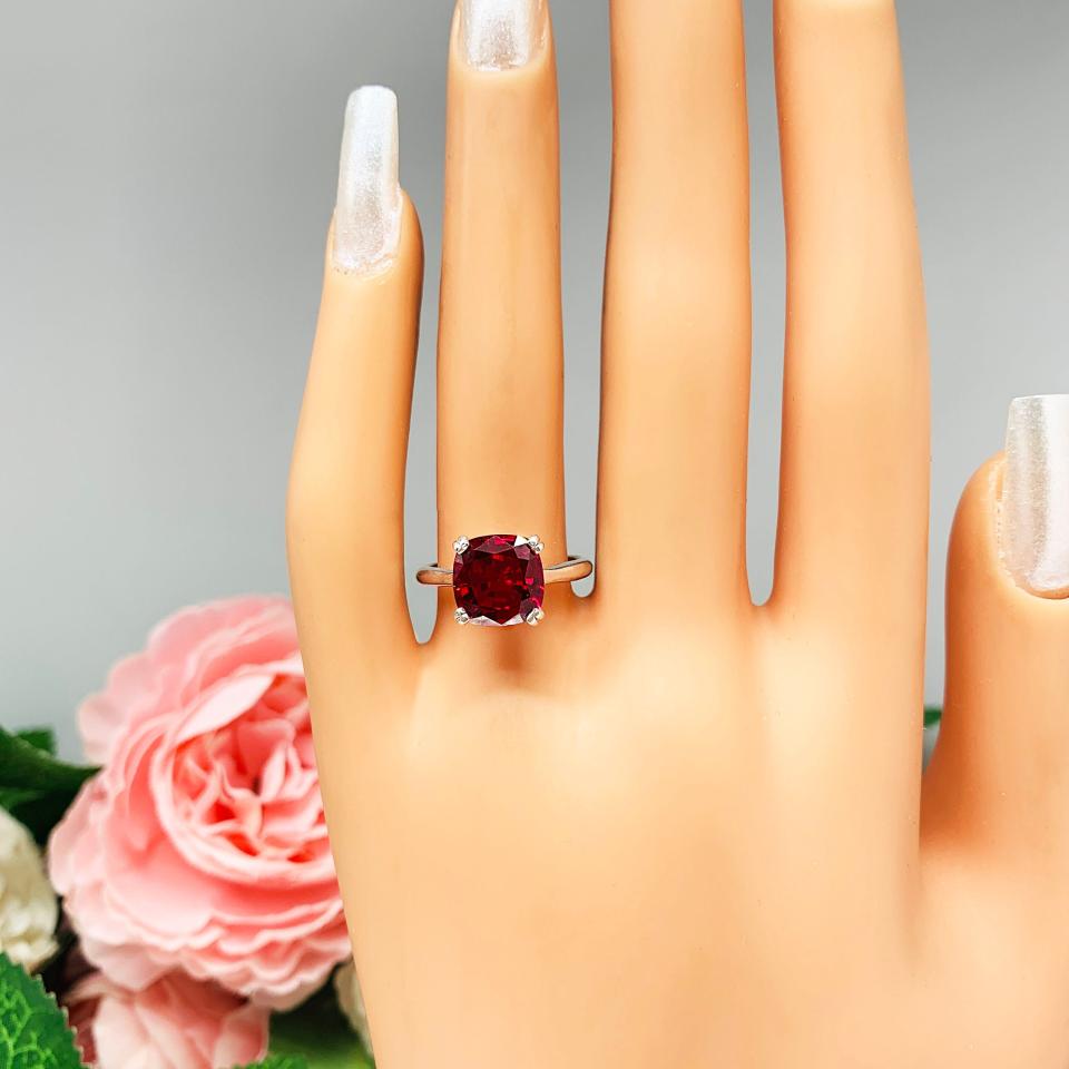 Mannequin hand with long white shimmery nails, displayed against a soft white-gray background with pink and white floral accents. The hand is wearing a sterling silver ring with a cushion cut lab created deep red ruby set in a split-prong setting.