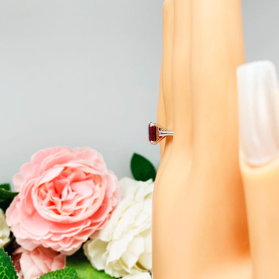 Mannequin hand with long white shimmery nails, displayed against a soft white-gray background with pink and white floral accents. The hand is wearing a sterling silver ring with a cushion cut lab created deep red ruby set in a split-prong setting.