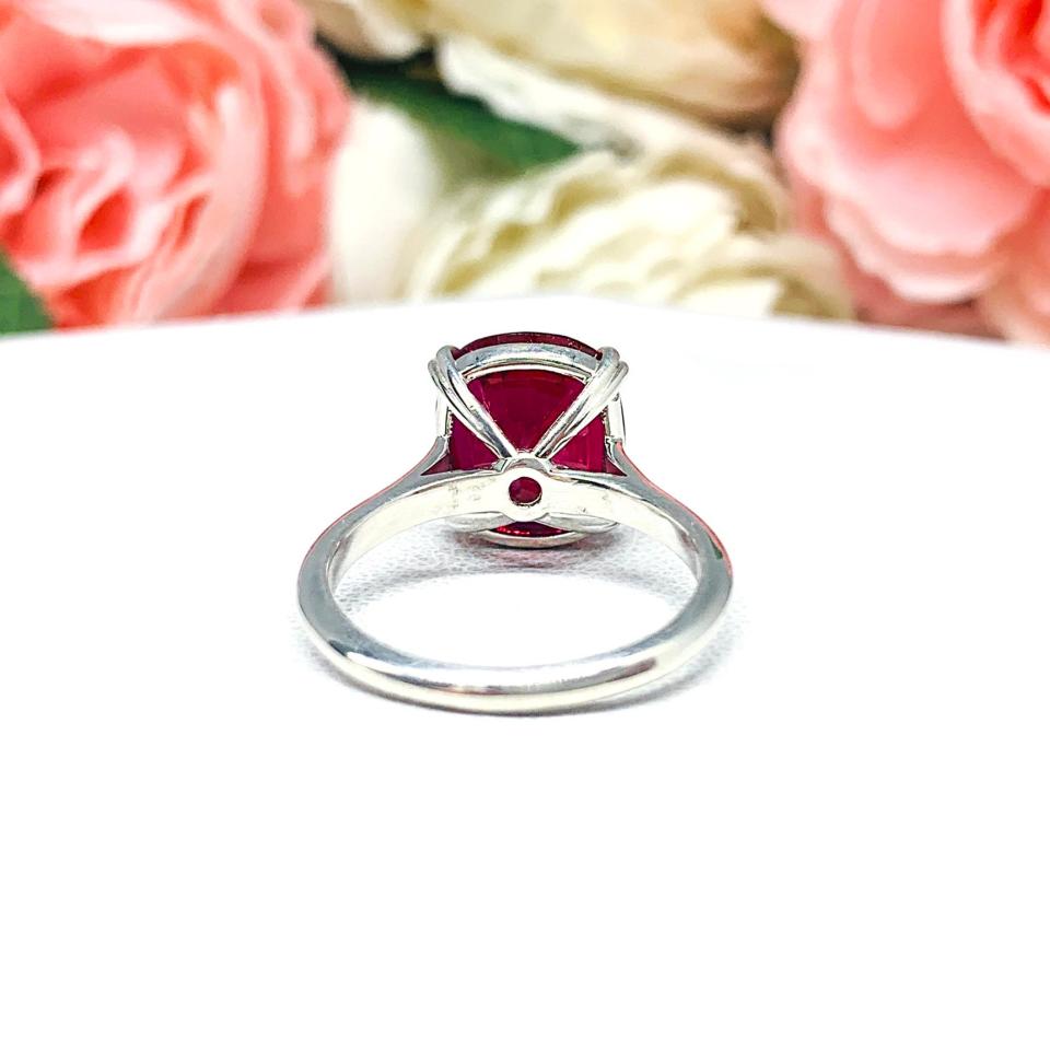 Sterling silver ring featuring a vivid elongated cushion-cut lab created ruby held in a split-prong setting, with soft-focus pink and white flowers in the background and “SJ” initials subtly visible in the corner.