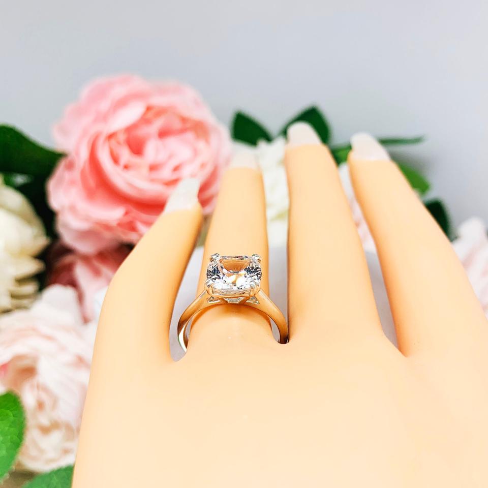 Mannequin hand with long white shimmery nails, displayed against a soft white-gray background with pink and white floral accents. The hand is wearing a sterling silver ring with a sparkling cushion cut lab created white sapphire set in a split-prong setting.