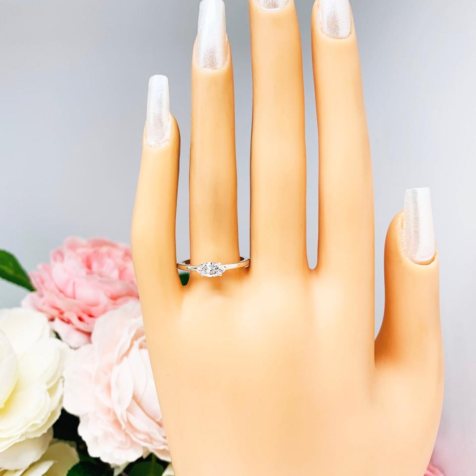 Mannequin hand with long white shimmery nails, displayed against a soft white-gray background with pink and white floral accents. The hand is wearing a dainty 4-prong sterling silver, horizontal east west set engagement ring featuring a precision-faceted marquise cubic zirconia.