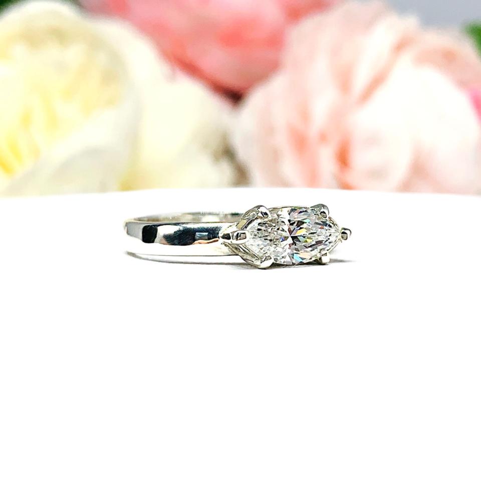 A 6-prong sterling silver, horizontal east west set engagement ring featuring a precision-faceted near colorless (G/H color) marquise cubic zirconia.  Displayed on a white surface with soft pastel floral background.