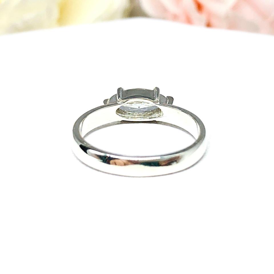 A 6-prong sterling silver, horizontal east west set engagement ring featuring a precision-faceted near colorless (G/H color) marquise cubic zirconia.  Displayed on a white surface with soft pastel floral background.