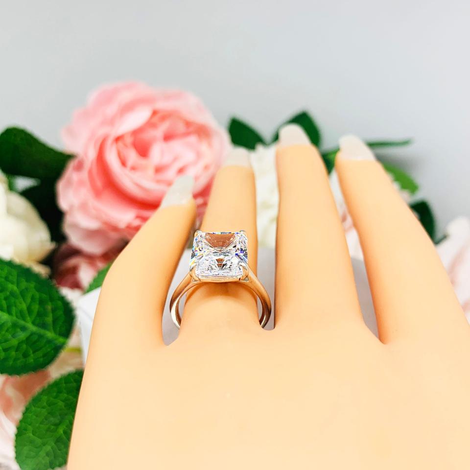 Mannequin hand with long white shimmery nails, displayed against a soft white-gray background with pink and white floral accents. The hand is wearing an elegant sterling silver cathedral engagement ring set with a large radiant cut cubic zirconia.