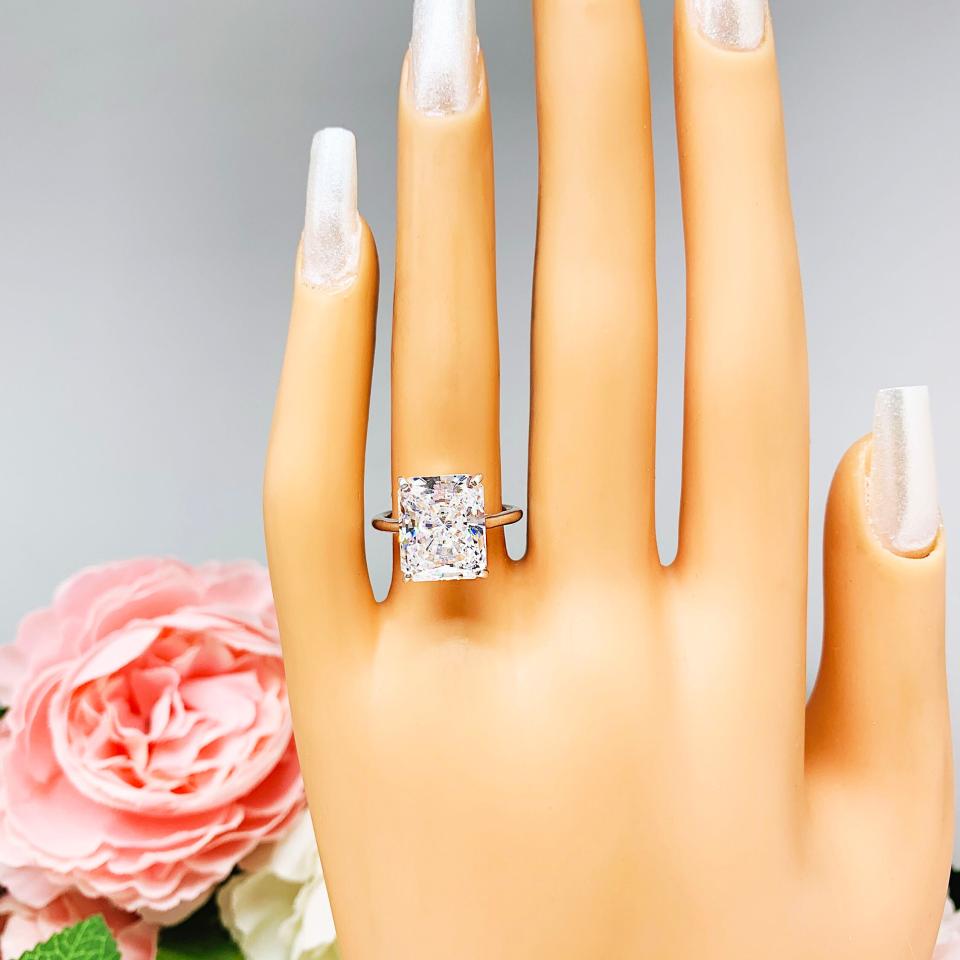 Mannequin hand with long white shimmery nails, displayed against a soft white-gray background with pink and white floral accents. The hand is wearing an elegant sterling silver cathedral engagement ring set with a large radiant cut cubic zirconia.