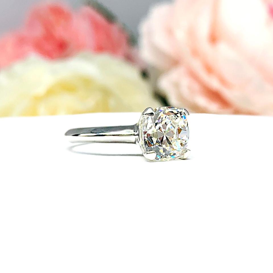 Sterling silver engagement ring set with an 8mm old mine cut near colorless cubic zirconia. The ring is resting on a white surface with a background of soft-focus pink and white flowers.