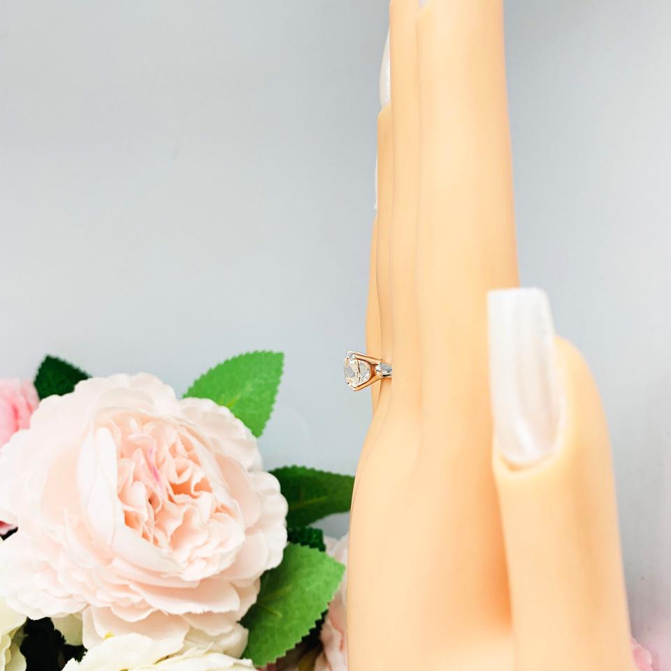 Mannequin hand with long white shimmery nails, displayed against a soft white-gray background with pink and white floral accents. The hand is wearing a sterling silver engagement ring set with an 8mm old mine cut near colorless cubic zirconia.