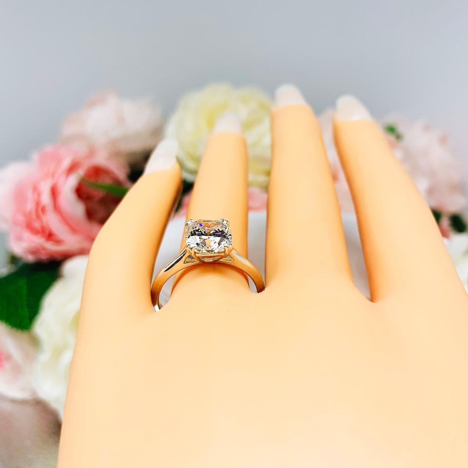 A display hand with long white fingernails standing up and with the top of the hand facing forward, displayed against a soft-focus background of pink and white flowers. The display hand is wearing sterling silver cathedral-style engagement ring featuring an 8mm near colorless cushion cut cubic zirconia center stone.