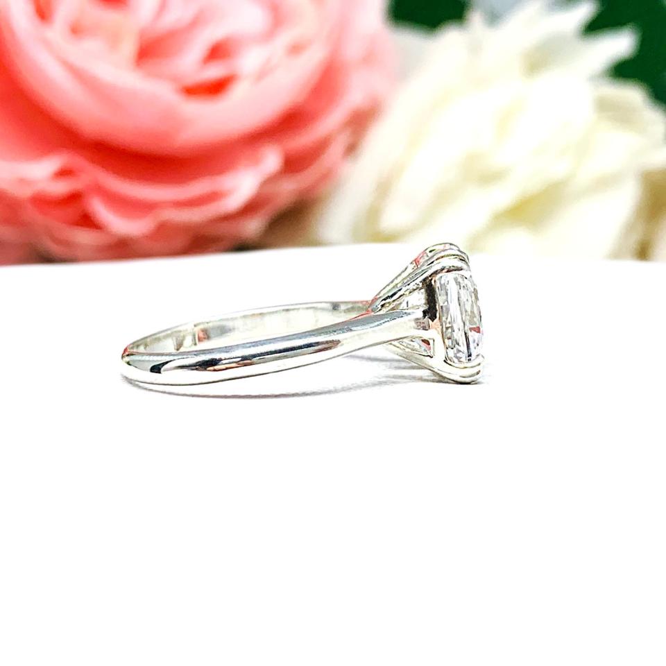 Sterling silver engagement ring with a sparkling cushion cut lab created white sapphire set in a split-prong setting set against a white surface with soft-focus pink and white flowers in the background.