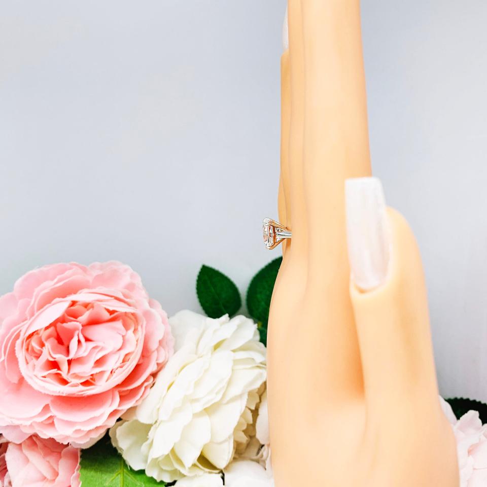 Mannequin hand with long white shimmery nails, displayed against a soft white-gray background with pink and white floral accents. The hand is wearing a sterling silver ring with a sparkling cushion cut lab created white sapphire set in a split-prong setting..