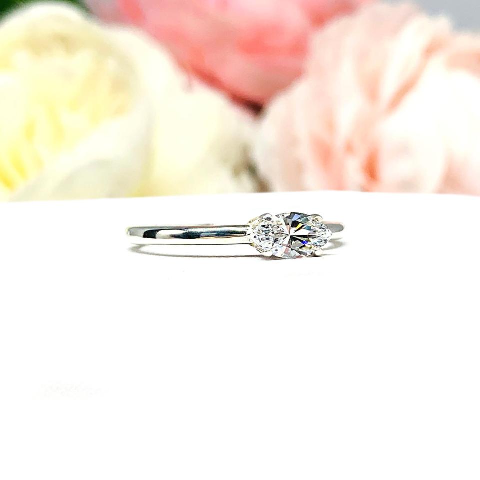 Dainty 4-prong sterling silver, horizontal east west set engagement ring featuring a precision-faceted marquise cubic zirconia.  Displayed on a white surface with soft pastel floral background.