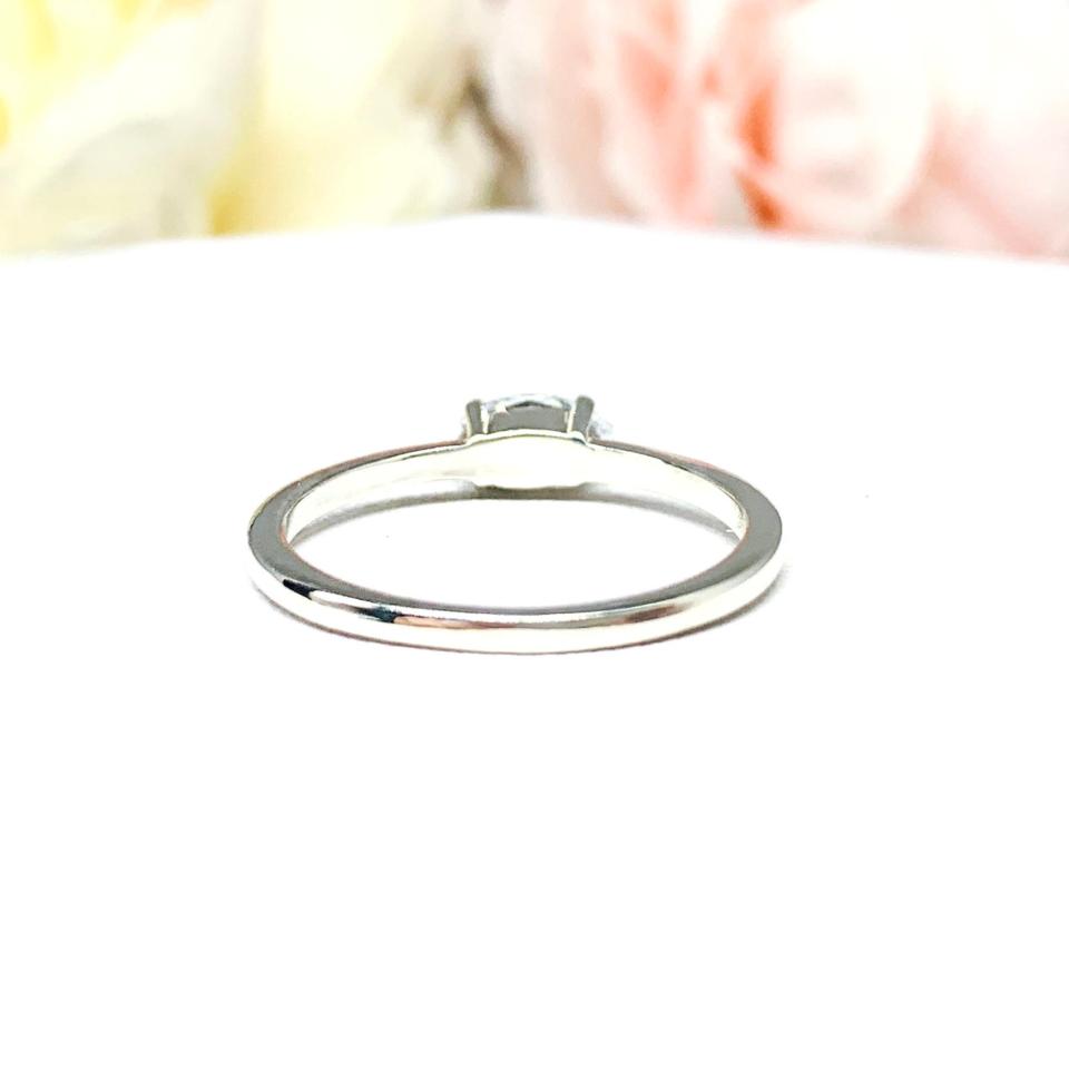 Dainty 4-prong sterling silver, horizontal east west set engagement ring featuring a precision-faceted marquise cubic zirconia.  Displayed on a white surface with soft pastel floral background.