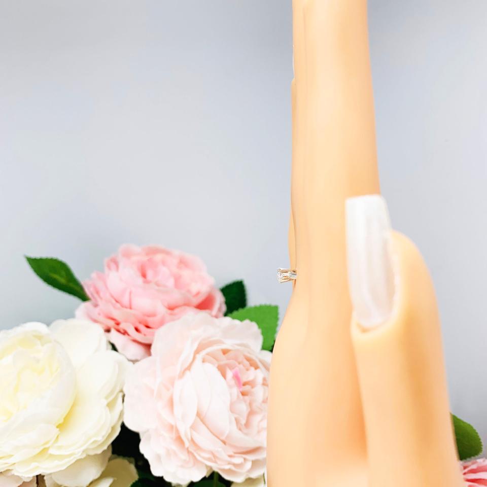 Mannequin hand with long white shimmery nails, displayed against a soft white-gray background with pink and white floral accents. The hand is wearing a dainty 4-prong sterling silver, horizontal east west set engagement ring featuring a precision-faceted marquise cubic zirconia.