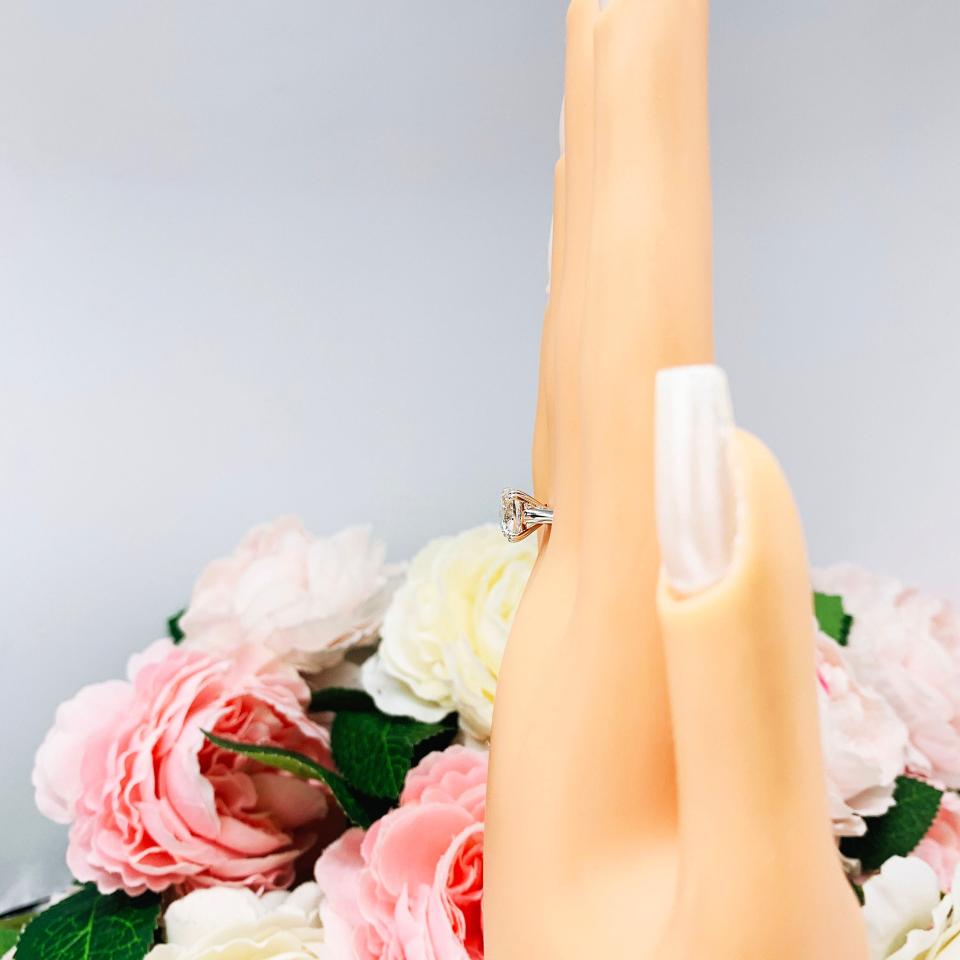 A display hand with long white fingernails standing up and with the top of the hand facing forward, displayed against a soft-focus background of pink and white flowers. The display hand is wearing sterling silver cathedral-style engagement ring featuring an 8mm near colorless cushion cut cubic zirconia center stone.