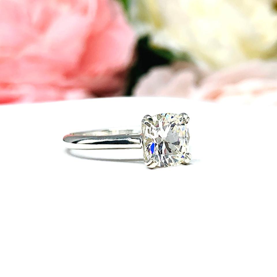 Sterling silver split prong cathedral engagement ring set with an 7.5mm near colorless old mine cut cubic zirconia. The ring is resting on a white surface with a background of pink and white flowers.