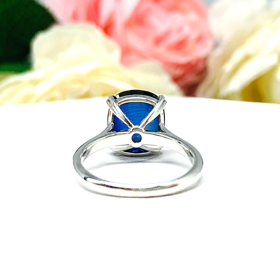 Sterling silver ring featuring a vivid elongated cushion-cut lab created blue sapphire held in a split-prong setting, with soft-focus pink and white flowers in the background.