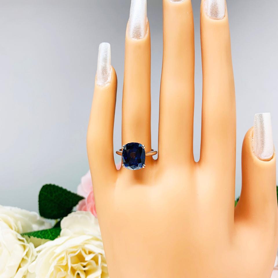 Mannequin hand with long white shimmery nails, displayed against a soft white-gray background with pink and white floral accents. The hand is wearing a sterling silver ring featuring a vivid elongated cushion-cut lab created blue sapphire held in a split-prong setting.