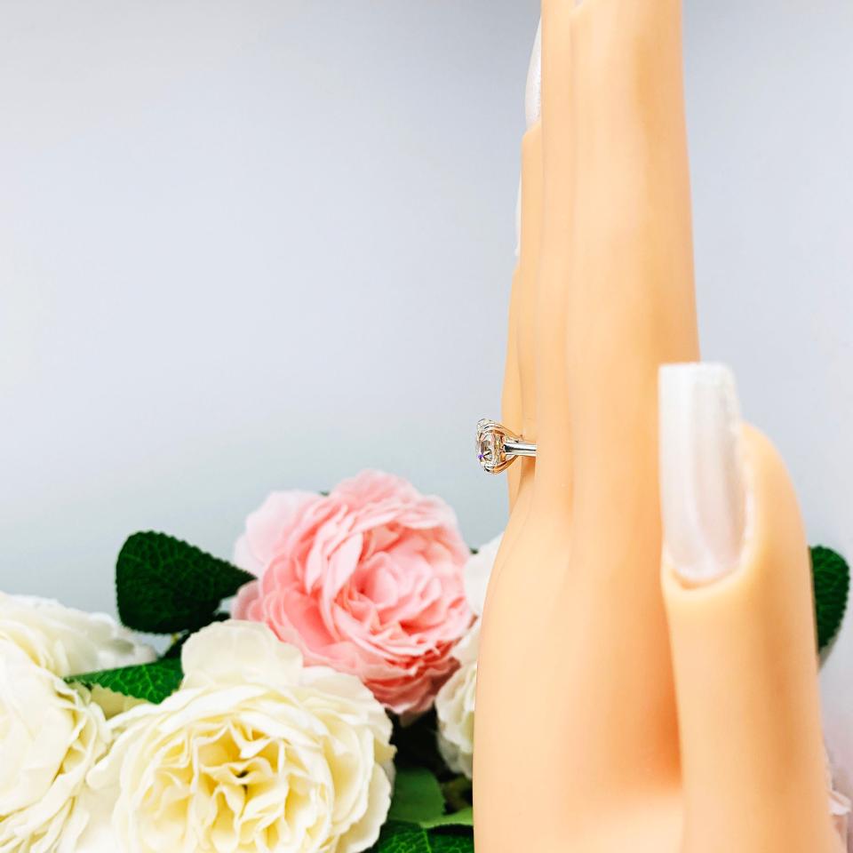 A display hand with long white fingernails standing up and with the top of the hand facing forward, displayed against a soft-focus background of pink and white flowers. The display hand is wearing a 9mm (3 ct) old European cut near-colorless CZ solitaire engagement ring. Set in a split prong cathedral-style mount, the ring gleams with vintage-inspired elegance.
