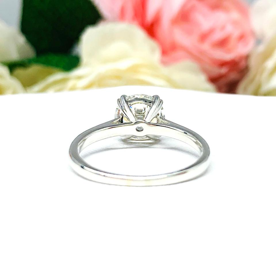 A romantic close-up of a 9mm (3 ct) old European cut near-colorless CZ solitaire engagement ring. Set in a sterling silver split prong cathedral-style mount, the ring gleams with vintage-inspired elegance. Soft pink and white flowers blur in the background, adding a dreamy, bridal ambiance.