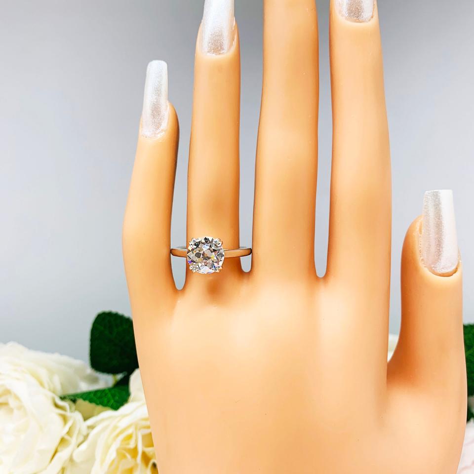 A display hand with long white fingernails standing up and with the top of the hand facing forward, displayed against a soft-focus background of pink and white flowers. The display hand is wearing a 9mm (3 ct) old European cut near-colorless CZ solitaire engagement ring. Set in a split prong cathedral-style mount, the ring gleams with vintage-inspired elegance.