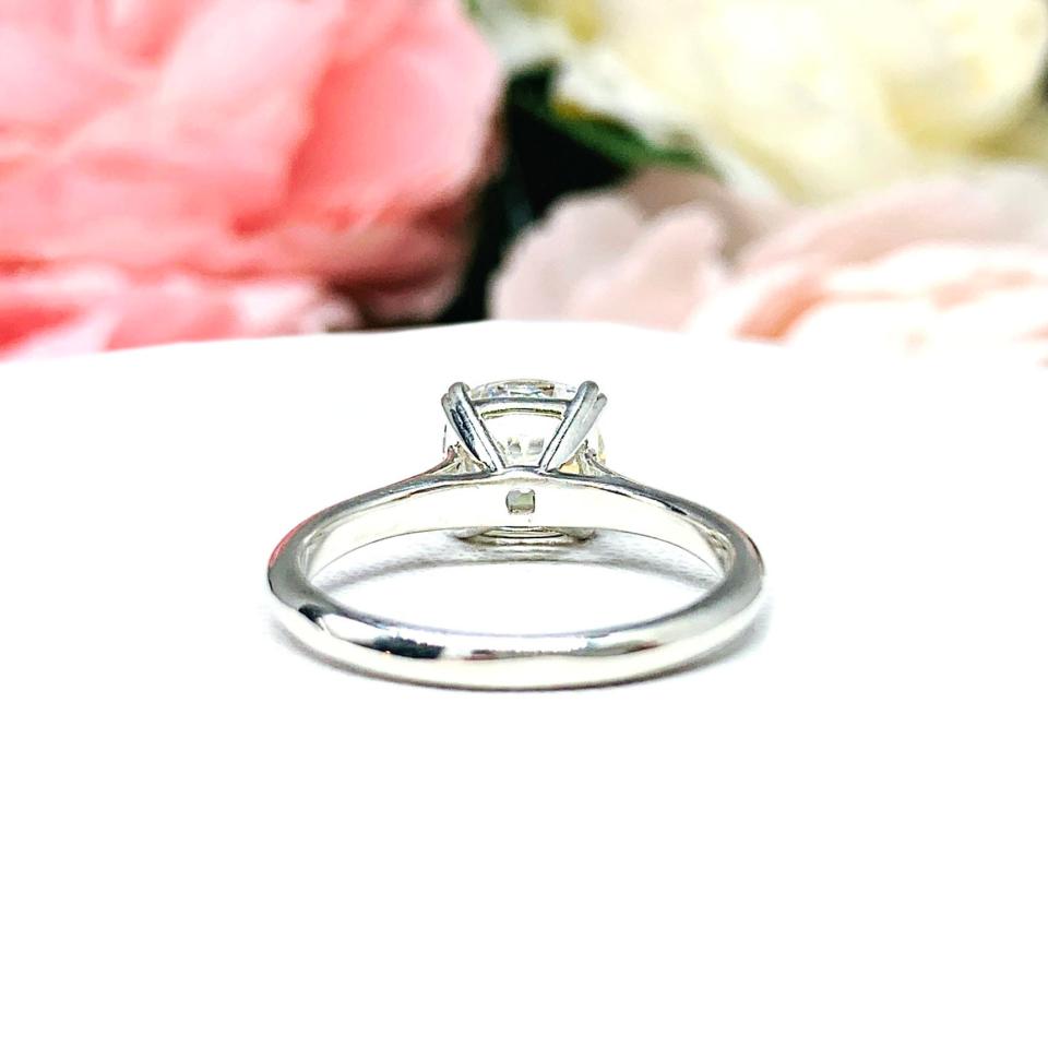 Sterling silver split prong cathedral engagement ring set with an 7.5mm near colorless old mine cut cubic zirconia. The ring is resting on a white surface with a background of pink and white flowers.