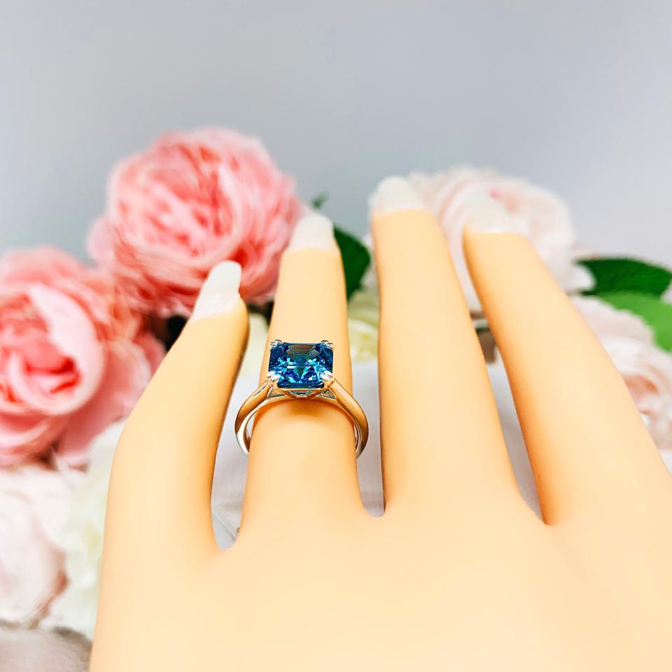 Mannequin hand with long white shimmery nails, displayed against a soft white-gray background with pink and white floral accents. The hand is wearing a sterling silver 8mm teal blue asscher cut cubic zirconia solitaire ring.