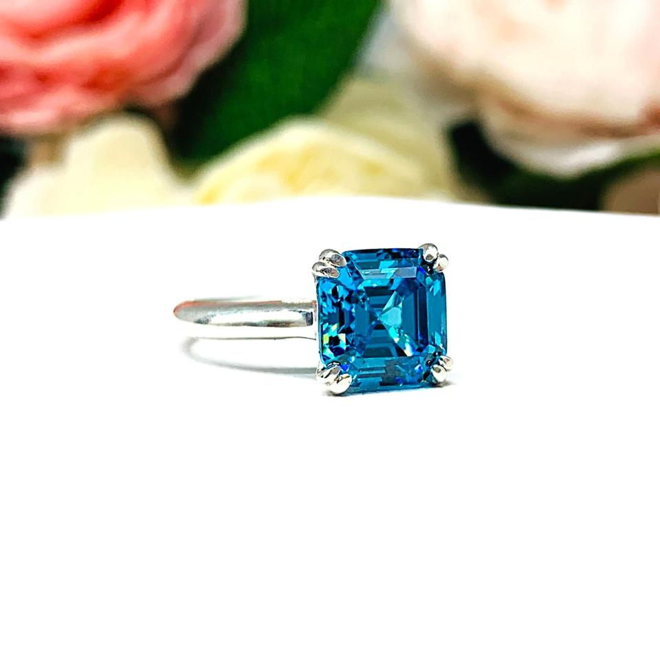 Sterling silver 8mm teal blue asscher cut cubic zirconia solitaire ring. The ring is resting on a white surface with a background of pink and white flowers.