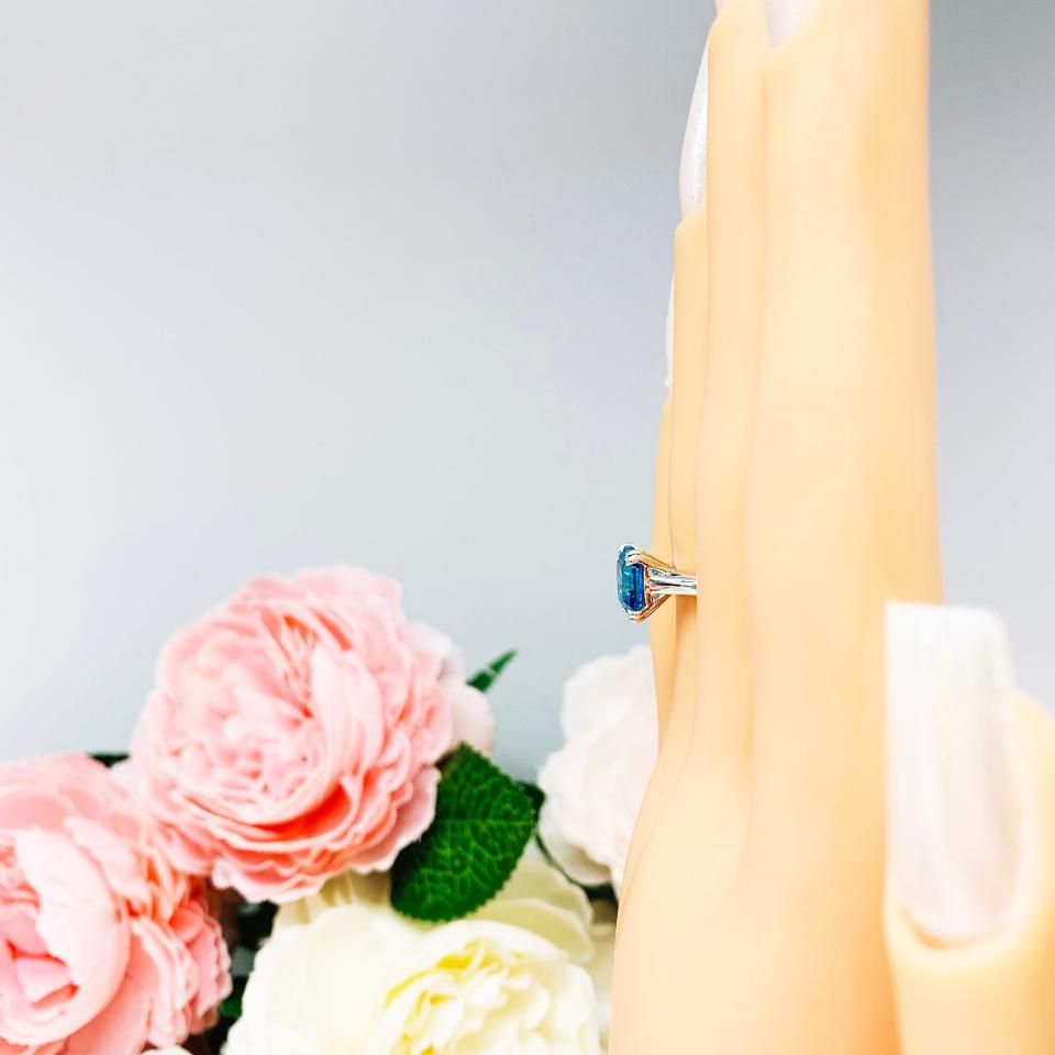 Mannequin hand with long white shimmery nails, displayed against a soft white-gray background with pink and white floral accents. The hand is wearing a sterling silver 8mm teal blue asscher cut cubic zirconia solitaire ring.