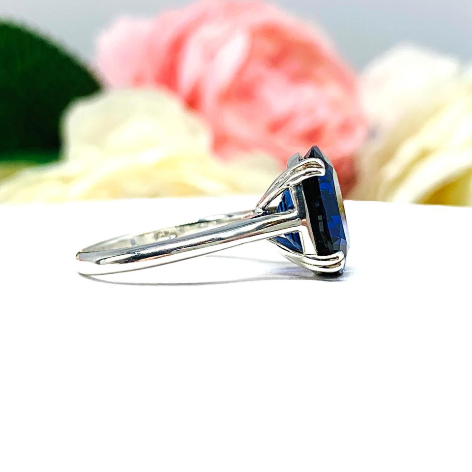 Sterling silver ring featuring a vivid elongated cushion-cut lab created blue sapphire held in a split-prong setting, with soft-focus pink and white flowers in the background.