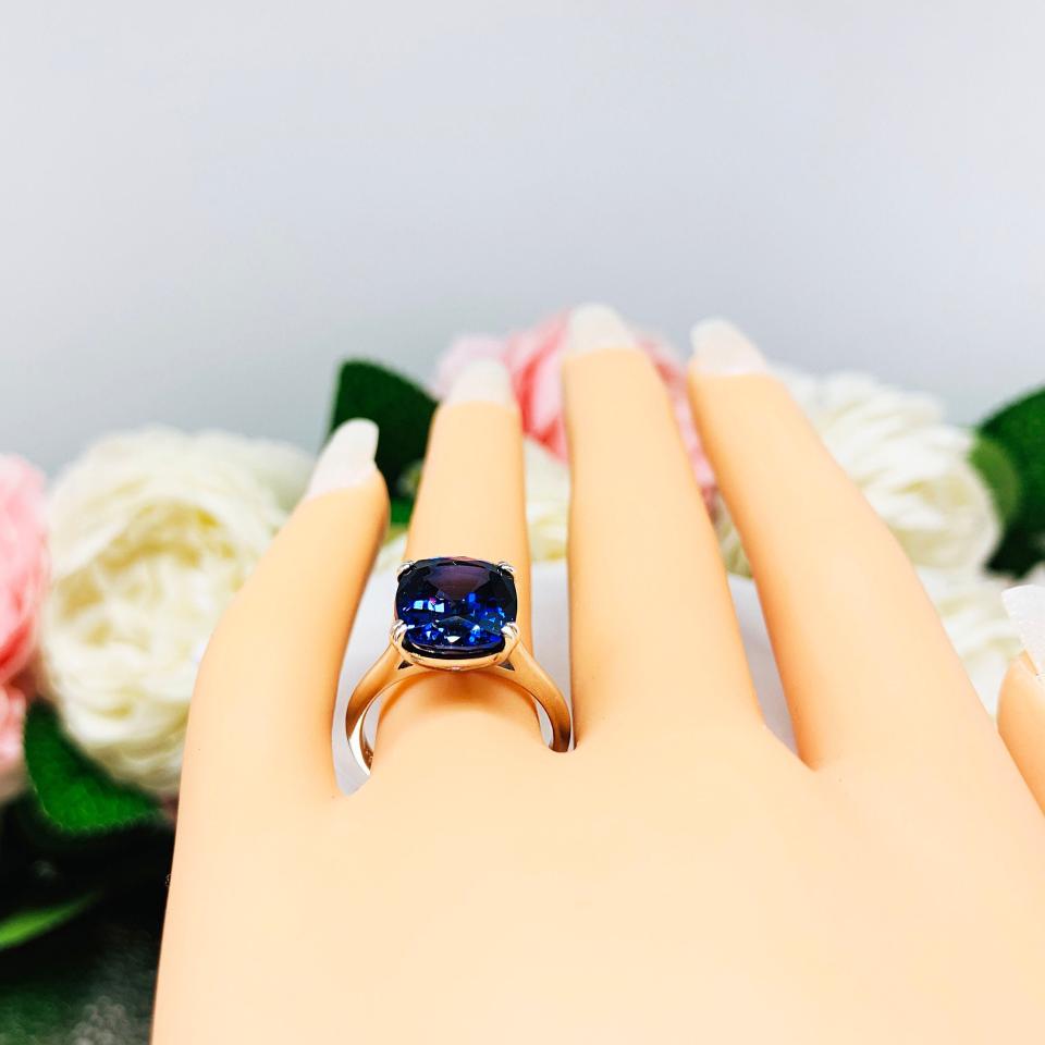 Mannequin hand with long white shimmery nails, displayed against a soft white-gray background with pink and white floral accents. The hand is wearing a sterling silver ring featuring a vivid elongated cushion-cut lab created blue sapphire held in a split-prong setting.