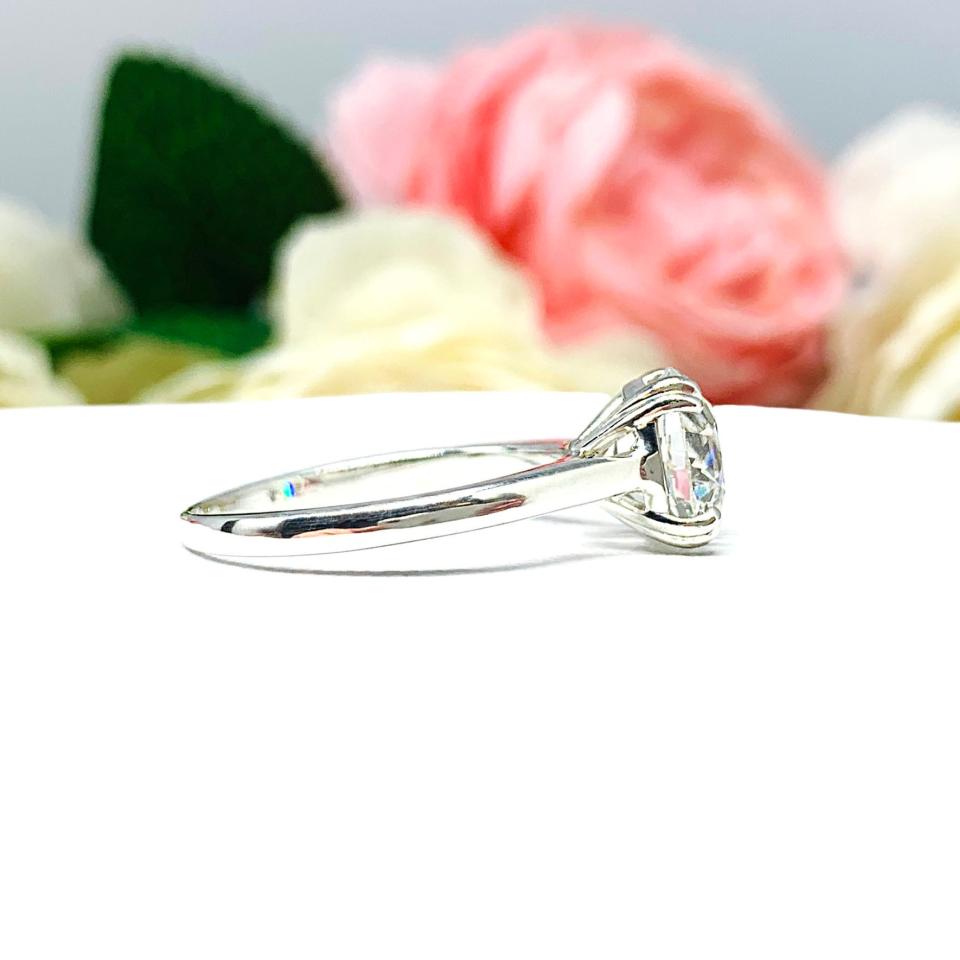 A romantic close-up of a 9mm (3 ct) old European cut near-colorless CZ solitaire engagement ring. Set in a sterling silver split prong cathedral-style mount, the ring gleams with vintage-inspired elegance. Soft pink and white flowers blur in the background, adding a dreamy, bridal ambiance.